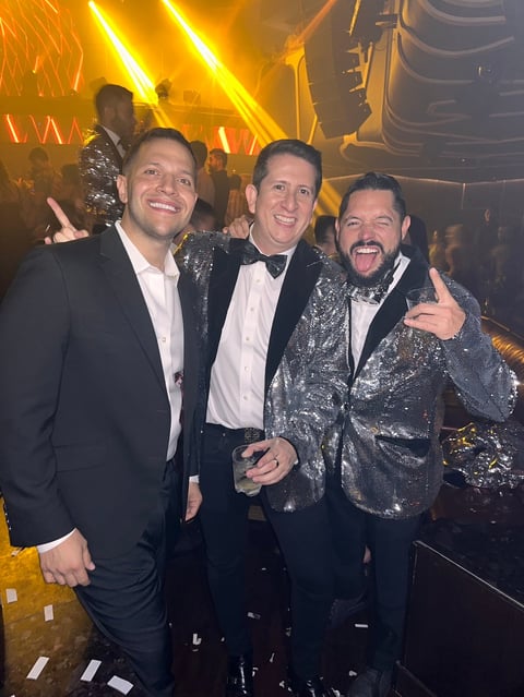 Three men posing at a nightclub celebration with gold neon lights and stage lighting in the background